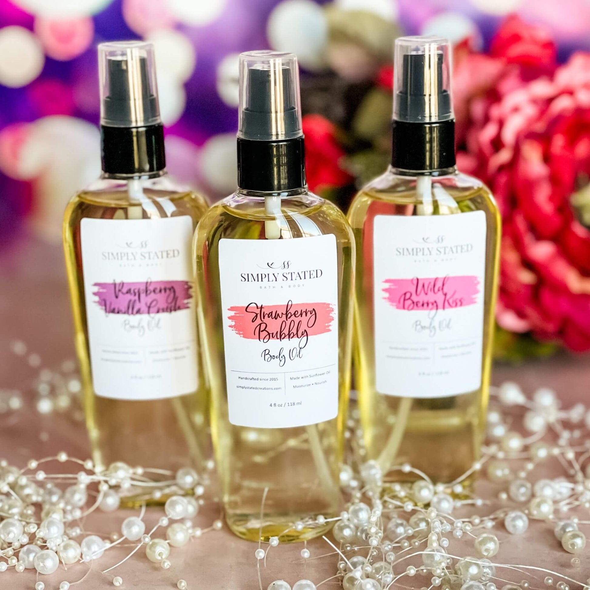 A trio of Sweet Temptations Body Oils—Raspberry Vanilla Crush, Strawberry Bubbly, and Wild Berry Kiss—arranged on a blush pink background with pearl accents and vibrant floral decor.